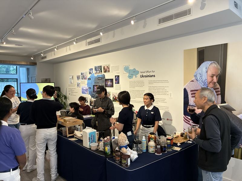 Students serving drinks and refreshments from a long table beside a wall display titled Relief Effort for Ukrainians, with visitors talking in the foreground.