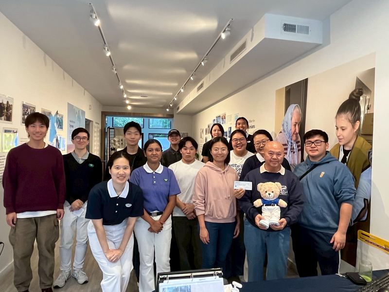 A group of about fifteen students gathered for a photo indoors, one holding a donation cheque and a Tzu Chi bear.