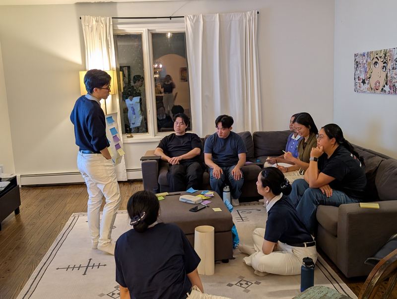 A student stands beside a wall of sticky notes, presenting to eight others seated on couches and the floor of a living room.