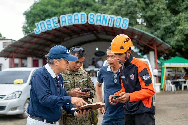 A Tzu Chi volunteer in a blue cap reviews notes with soldiers and a rescue worker in an orange helmet at a relief site in the Philippines.