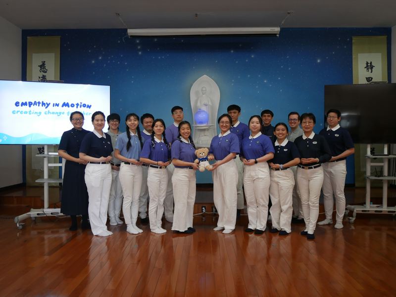 Around twenty students in matching purple and navy polos stand in a line, one holding a small Tzu Chi bear, in front of a screen reading Empathy in Motion.
